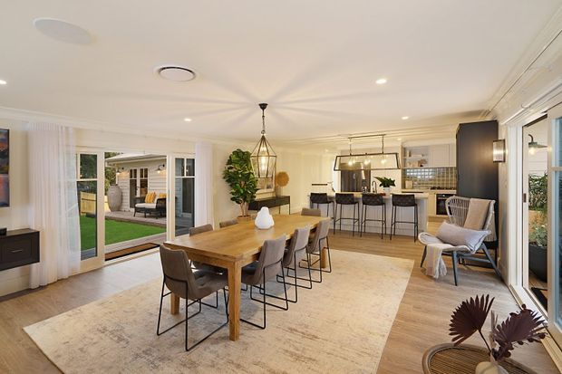 Bright open-plan dining and kitchen area with a large timber dining table, upholstered dining chairs, kitchen island with bar seating, pendant lighting, light timber flooring, and a neutral area rug. The room opens through extensive glazing and sliding doors to a small outdoor lawn and covered seating area. A lounge chair and indoor plants add warmth to the contemporary space.