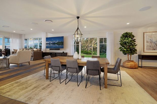 Bright open-plan dining and living area with a large timber dining table, upholstered dining chairs, a pale area rug, and a statement pendant light. Floor-to-ceiling windows and glazed doors provide abundant natural light, while a neutral palette, timber flooring, built-in cabinetry, artwork, and indoor planting create a refined contemporary setting.