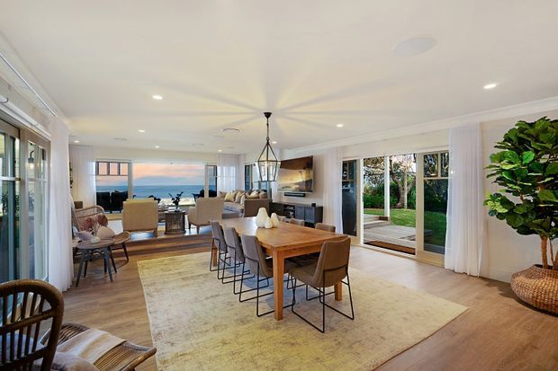 Bright open-plan coastal living and dining area with warm timber flooring, a large dining table, upholstered dining chairs, a lounge seating zone, and extensive floor-to-ceiling glazing opening to a lawn and ocean view. A linear pendant hangs over the dining table, while white walls and curtains create an airy, relaxed interior.