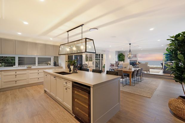 Bright open-plan coastal home interior featuring a large painted kitchen with central island, integrated cabinetry, pendant lighting, and timber-look flooring. The kitchen opens directly into a dining and living area with ocean views through glazing, creating an airy, relaxed entertaining space.