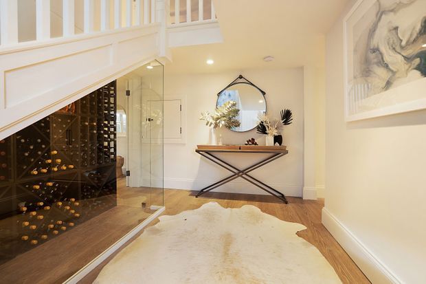 Refined residential hallway beneath a staircase, featuring a dramatic glass-fronted wine cellar, pale timber flooring, white architectural balustrading and trim, a console table with decorative objects, a round wall mirror, and a large neutral hide rug. The space is warmly lit and connects visually to a glass-enclosed area.