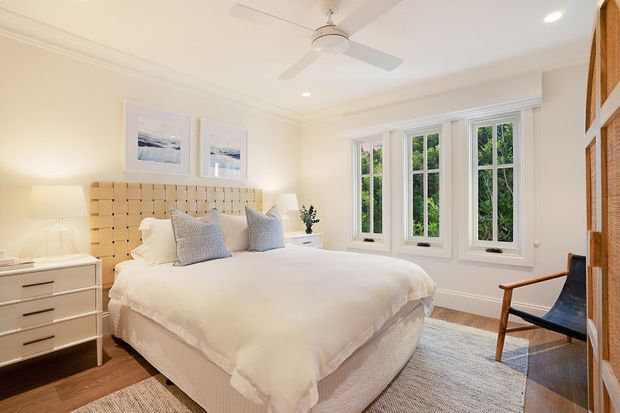 Bright, refined bedroom with a large upholstered bed, tufted beige headboard, layered white bedding, bedside tables, framed artwork, and a black leather accent chair. A bank of white-trimmed windows provides abundant natural light and views of greenery, while a ceiling fan, timber flooring, and neutral rug complete the room.