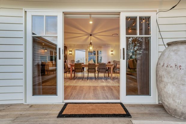 View from an exterior deck through open glazed doors into a warm, refined dining room. The room features a long timber dining table with upholstered chairs, pendant lighting, wall sconces, timber flooring, and extensive glazing. The exterior has pale horizontal weatherboard cladding and a large decorative ceramic vessel beside the entry.