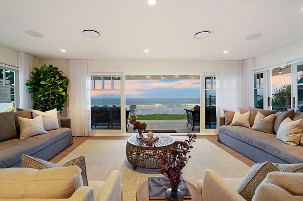 Bright coastal living room with symmetrical grey sofas and cream armchairs arranged around a round woven coffee table, opening through large glazed doors to a lawn and ocean view. White walls, sheer curtains, recessed lighting, and abundant windows create an airy, relaxed interior.