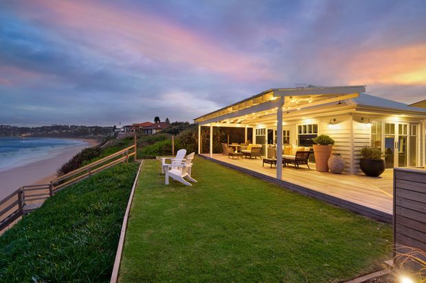 A warmly illuminated coastal home at dusk, viewed from its landscaped lawn and timber deck. The single-storey building features horizontal light-coloured weatherboard cladding, a broad low-pitched roof, extensive covered outdoor living, glazed doors and windows, and timber deck flooring. Outdoor seating and planters frame the deck, while a beach and coastal settlement are visible beyond the landscaped edge.