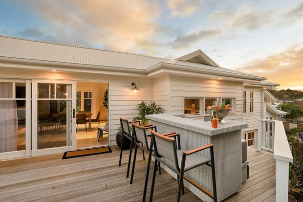 Elevated outdoor deck with a built-in tiled outdoor kitchen and breakfast bar, connected to a light weatherboard home. The space includes bar seating, a timber decking surface, sliding glass doors opening to an interior dining area, and a pitched roof with a small gable detail. Warm evening light creates an inviting residential setting.