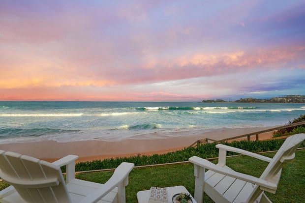 A tranquil beachfront scene viewed from a grassy seaside terrace, with white outdoor lounge chairs in the foreground overlooking sandy beach, turquoise ocean waves, and a rocky coastal headland under a pastel pink and blue sunset sky.