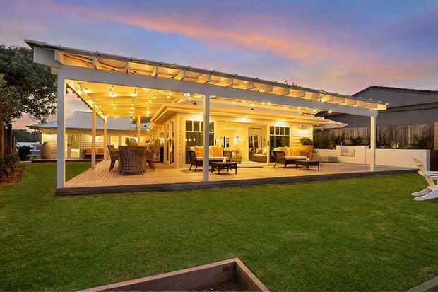 Evening exterior view of a contemporary single-storey home with a large timber deck and extensive covered outdoor entertaining area. The open-sided pergola has a flat, slatted roof structure with integrated warm lighting, enclosing outdoor lounge and dining furniture. Glazed doors connect the deck to the interior, with a broad lawn in the foreground and neighboring residential structures in the background.