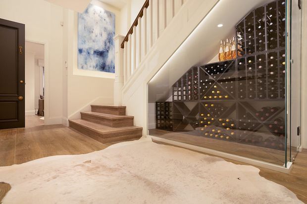 Elegant residential entryway with warm timber flooring, a short staircase, white paneled balustrade, dark front door, and a large glass-enclosed under-stair wine cellar with geometric bottle storage. A neutral hide rug and abstract artwork add softness and contrast.