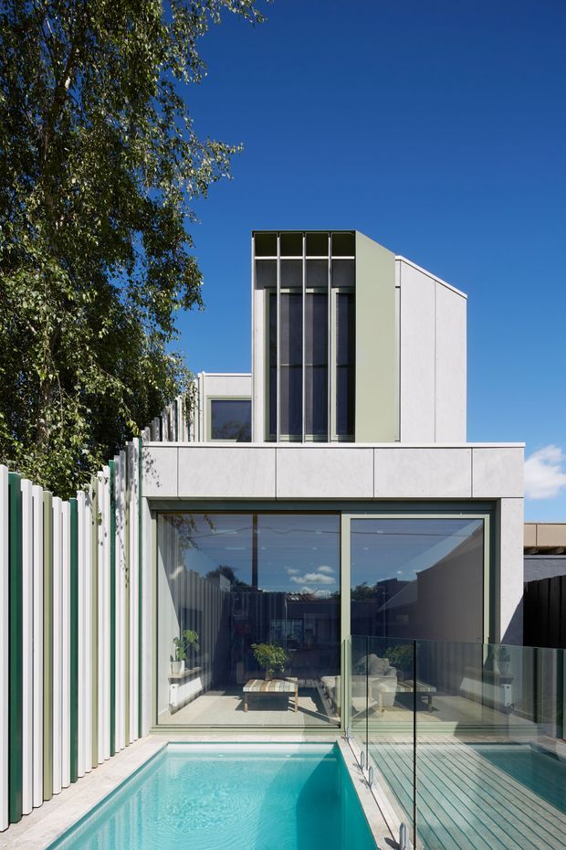 A contemporary multi-level urban residence viewed from the rear courtyard, with a compact turquoise swimming pool, timber deck, frameless glass balustrade, and large glazed living area. The upper storey features a sculptural pale rendered facade with tall vertical windows and projecting geometric volumes. A slatted privacy fence and mature tree border the property under a clear blue sky.