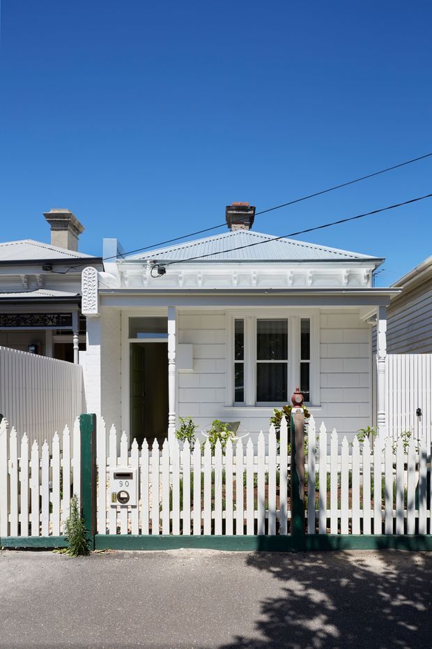 Front exterior view of a compact single-storey heritage-style cottage with white weatherboard cladding, a corrugated metal hipped roof, decorative veranda posts and fretwork, and a white picket fence. The house is presented in bright sunlight from the street, with neighbouring attached or closely spaced homes visible at either side.