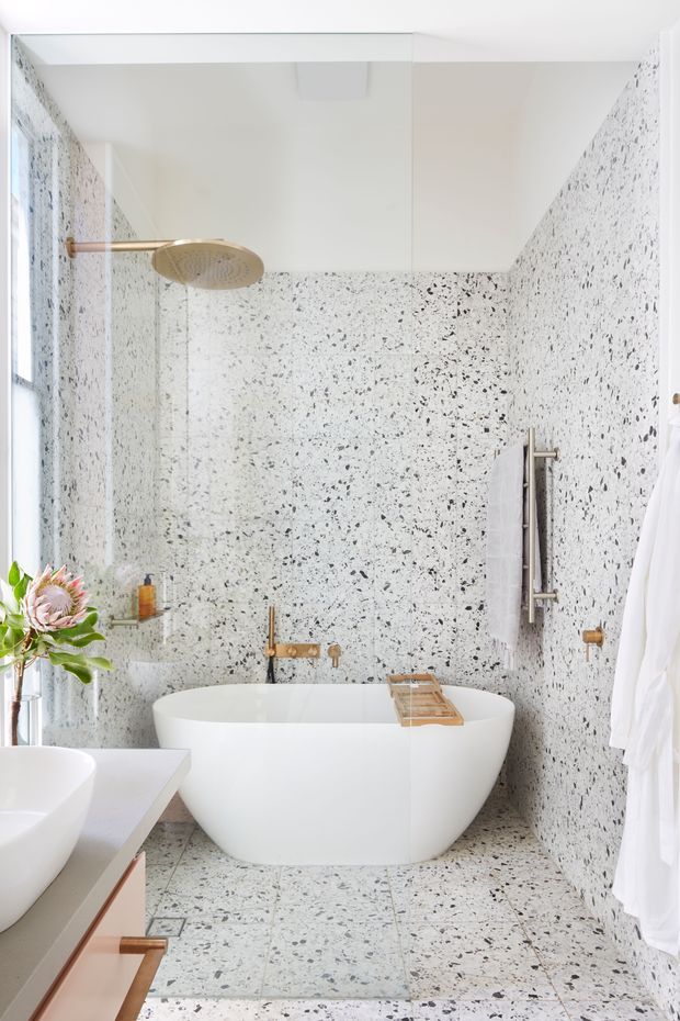 Bright contemporary bathroom featuring a freestanding white bathtub, terrazzo wall and floor surfaces with dark aggregate flecks, clear glass shower enclosure, and brushed brass shower and tapware. A partial white vanity is visible in the foreground, with a towel rail and white textiles on the right.