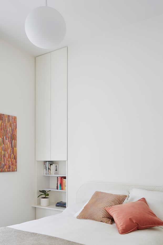 Bright, minimalist bedroom with a white upholstered bed, white bedding, and coral and terracotta accent pillows. A tall built-in white storage and open shelving unit occupies the corner, holding books and a small potted plant. The room has pale walls, a large spherical pendant light, and a colourful abstract artwork partially visible on the left wall.