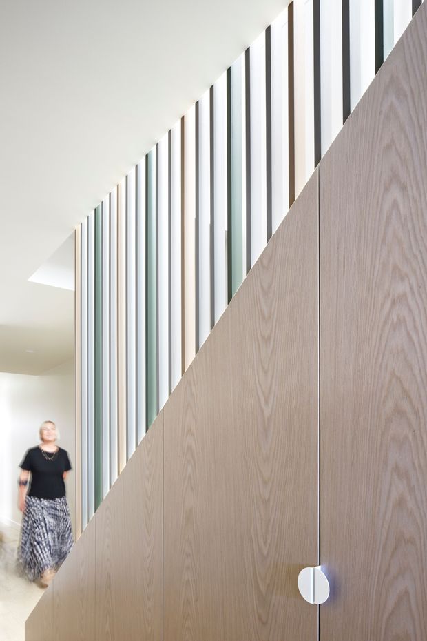 A bright contemporary interior hallway or stairway featuring a tall vertical glazed screen with alternating slim timber and dark framing elements. A large diagonal plane of light timber veneer cabinetry or wall panelling dominates the foreground, with a minimal white circular handle. The space has a clean white ceiling, pale flooring, and a person visible in the background for scale.