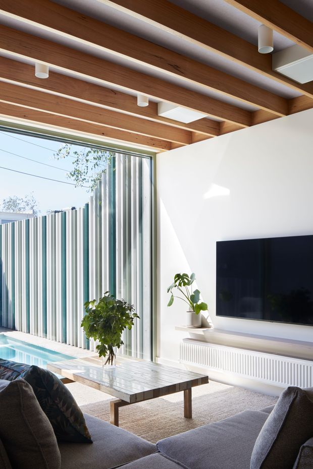 Bright contemporary living room with a large sectional sofa facing a wall-mounted television and slim built-in console. The room features a dramatic exposed timber beam ceiling with integrated cylindrical and linear lighting, white walls, a pale textured rug, and a marble-topped coffee table. Full-height glazing opens toward a narrow swimming pool and a vertically striped privacy screen outside, bringing in abundant natural light.