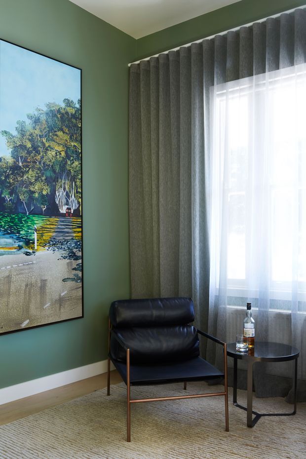 A refined sitting corner with deep sage-green walls, a large framed landscape artwork, full-height grey textured curtains with sheer panels, and a black leather lounge chair with a slim bronze-toned metal frame. A small round black side table holds a bottle, and a neutral textured rug covers the floor. The room has strong natural light and a restrained, sophisticated palette.