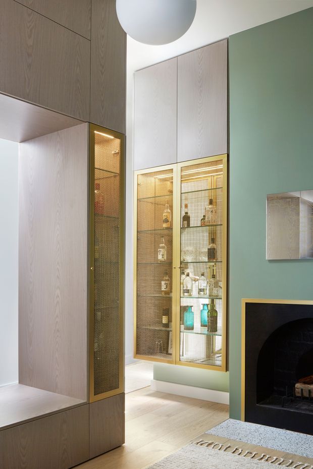 A refined contemporary living room featuring extensive light timber built-in cabinetry, a tall illuminated glass-fronted display cabinet with brass-toned mesh and framing, and a muted sage-green feature wall. A black fireplace with a gold-toned trim occupies the lower right, while a large spherical pendant light and pale timber flooring complete the warm, elegant interior.