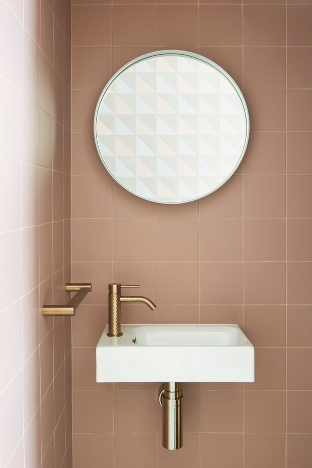 Compact bathroom with a wall-mounted white rectangular basin, exposed chrome drainpipe, brushed brass tapware, and a round framed mirror. The walls are clad in dusty pink-brown square tiles with matching grout, while the mirror has a subtle geometric pale pattern.