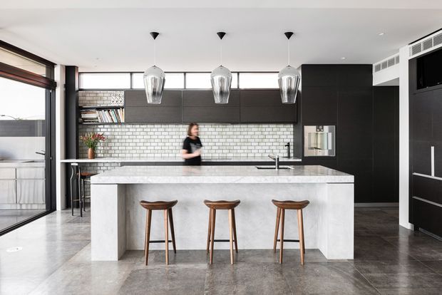 Contemporary kitchen featuring a large white marble-look island with seating for three, dark built-in cabinetry, a white subway-tile splashback, timber bar stools, and three pendant lights. Industrial-style glazing and polished grey flooring contribute to a monochromatic, refined interior.