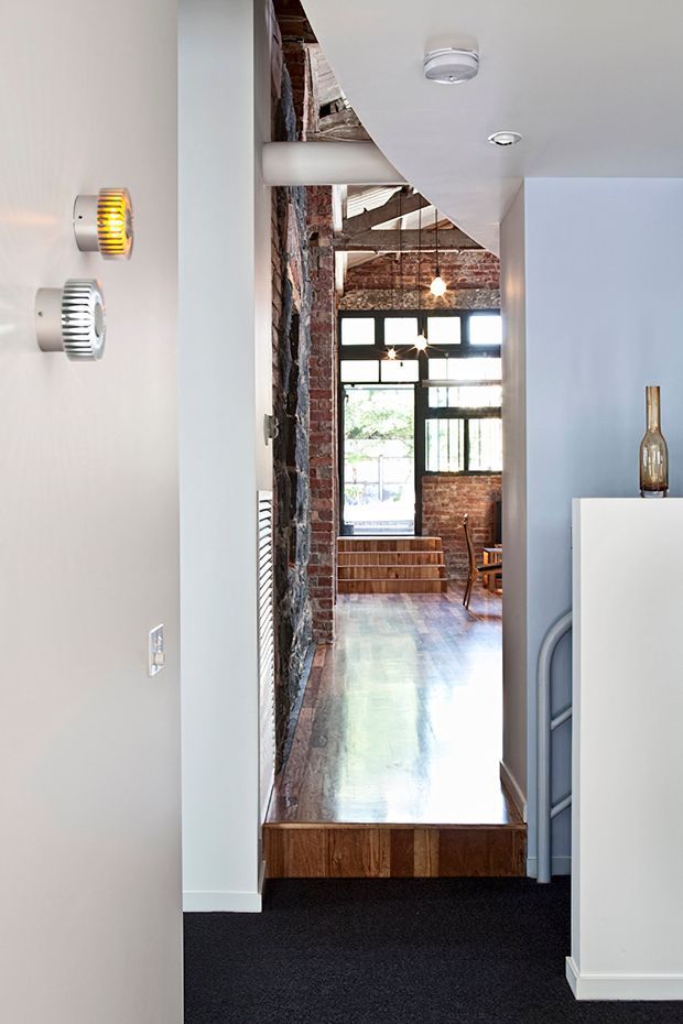 Interior view through a narrow hallway into a loft-like living or dining space. The foreground has pale painted walls, dark carpet, modern wall lights, and a curved ceiling transition. Beyond the threshold are exposed red brick walls, timber ceiling structure, large black-framed industrial-style glazing, polished timber flooring, timber steps, and a small dining area. The image combines contemporary finishes with original industrial architectural fabric.