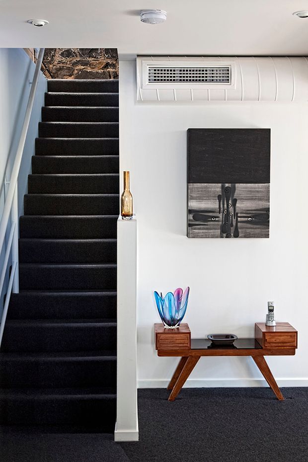A compact interior hallway and stairway with a dark charcoal carpeted staircase, white walls and ceiling, and a mid-century-style timber console table. Colourful glass decor, a small sculpture, artwork, and a wall-mounted air-conditioning unit are visible. The stairwell leads upward toward a stone-textured wall or landing.