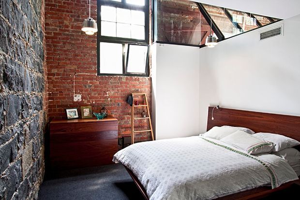 Industrial-style bedroom with an exposed red-brick wall, dark timber bed and dresser, black-framed factory windows, exposed structural elements, and a white bedding scheme. A textured stone-like feature wall and dark carpet add contrast, while pendant lights and a leaning timber ladder contribute to the loft aesthetic.