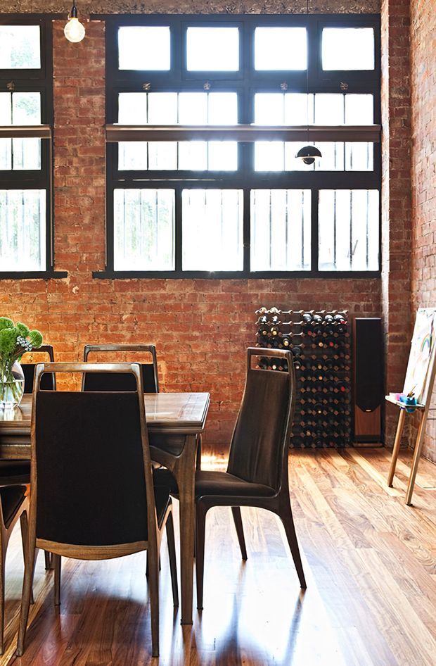 A sunlit industrial-style dining area with exposed red brick walls, tall black-framed multi-pane windows, polished timber flooring, and a dark timber dining table surrounded by black upholstered chairs. A freestanding wine rack, floor-standing speaker, pendant lights, and an easel occupy the perimeter, creating a warm loft-like atmosphere.