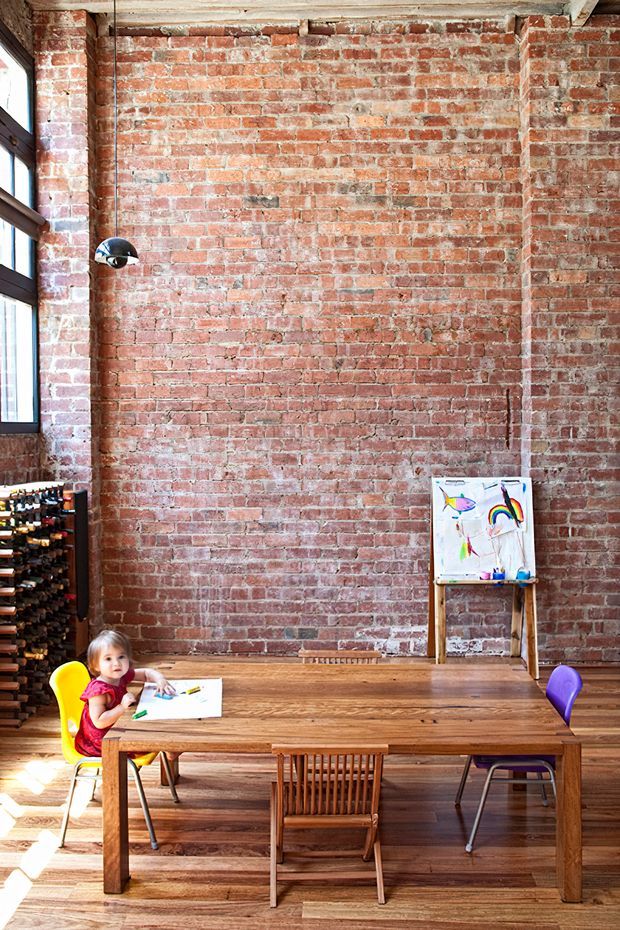 A tall, loft-like interior with exposed red brick walls, timber flooring, and a large solid timber dining or activity table. Dark timber-framed windows line the left side, while a wall-mounted pendant light, children's chairs, artwork, and an easel create a casual creative workspace or family dining setting. The room has an industrial character softened by warm wood and natural daylight.