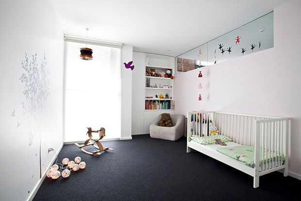 Bright, playful nursery with a white cot, dark carpet, built-in open shelving filled with children's books and toys, a small upholstered armchair, rocking horse, hanging pendant light, and whimsical wall decals. A large window provides soft natural light, while a mirrored upper wall reflects the room and decorations.