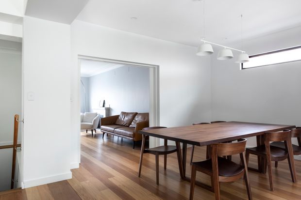 Bright, minimalist dining area with a large dark timber dining table and matching wooden chairs. White walls and ceiling, timber flooring, a linear arrangement of white pendant lights, and a high horizontal window create an airy contemporary interior. An adjoining living room with a leather sofa and armchair is visible through the wide opening.