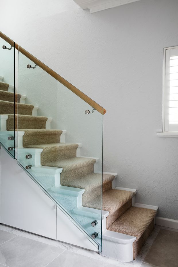 Interior view of a residential stairway with carpeted treads, white-painted stringers and risers, a clear glass balustrade secured with metal fittings, and a timber handrail. The surrounding walls and trim are white, with a shuttered window at right and pale tiled flooring below.