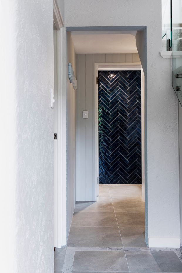 A narrow, bright interior passageway viewed through a doorway, with pale textured and vertical-panel walls, grey trim, and large light stone-look floor tiles. A dark blue-black glossy herringbone tile feature wall is visible at the far end, framed in white. The space has a clean, refined coastal-influenced look with strong contrast between the light corridor and dark tiled feature.