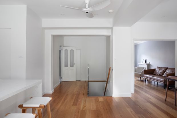 Bright white open-plan interior showing a hallway and stair opening, with a partial kitchen peninsula and stool in the foreground and a living room visible through the right-hand opening. The space has warm timber flooring, white walls and ceiling, a white ceiling fan, and simple contemporary furnishings.