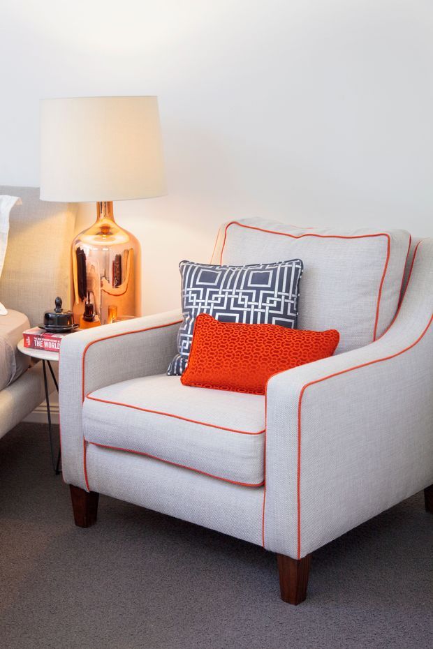 A close-up interior vignette of a light grey upholstered armchair with red piping and dark wooden legs, styled with geometric navy and white and textured red cushions. A matching sofa edge, side table, reflective amber table lamp, and neutral wall and carpet create a polished, colourful seating corner.
