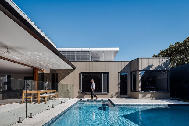 Contemporary single-storey residence centred around a bright swimming pool courtyard. The home features flat rooflines, expansive glazing, vertical timber-toned wall cladding, a covered outdoor entertaining area, and a clean white upper volume beneath a clear blue sky.