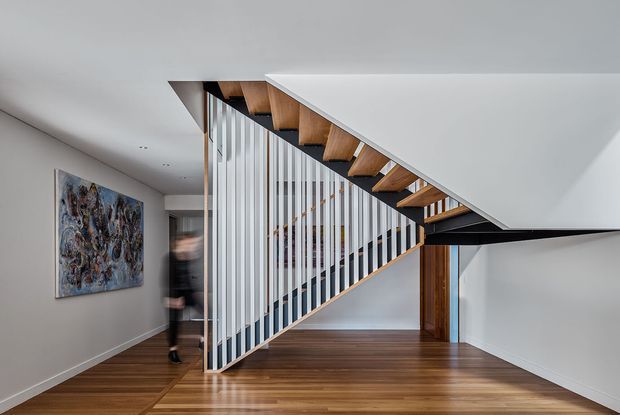 Contemporary interior hallway and stairway with a dramatic open-riser timber staircase, white vertical balustrade, dark structural stringer, polished timber flooring, and clean white walls. An abstract artwork and a partially visible glass doorway add visual interest.