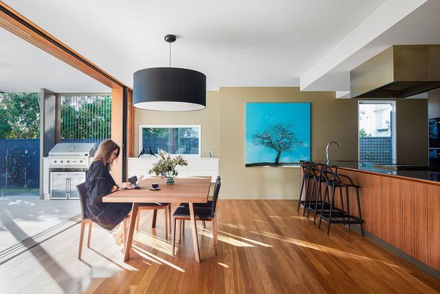 Bright open-plan kitchen and dining area with warm timber flooring and a timber dining table, opening to an outdoor cooking area through large glazed doors. The kitchen features dark cabinetry, a wood-fronted island with bar seating, and a large black pendant light. A turquoise artwork provides a strong accent against neutral walls.