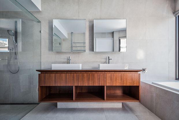 Contemporary bathroom featuring a wall-mounted double timber vanity with open shelving, two white vessel basins, twin mirrors, large-format grey wall and floor tiles, and a frameless glass walk-in shower. A bathtub is visible on the right beneath a window.