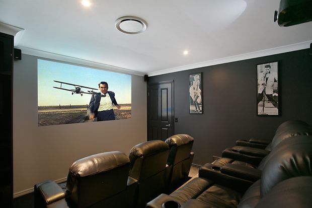 A dark, dedicated home cinema with multiple rows of large black leather reclining seats, a wall-mounted projection screen displaying a film, framed monochrome artwork, and recessed ceiling lighting. The room has dark painted walls, a white ceiling, and a compact enclosed layout.