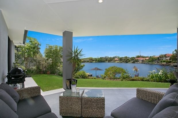 Covered outdoor living area overlooking a landscaped lawn and waterfront. The space features grey cushioned wicker lounge seating, a matching coffee table, concrete paving, a square structural column, ceiling downlights, and wide open views across the water to neighbouring homes.