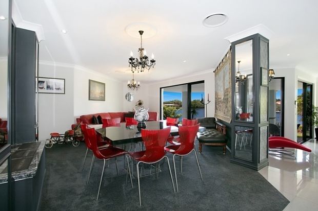 Bright, open-plan formal dining and living area with a large glass dining table surrounded by vivid red chairs, a red sectional sofa, dark carpet, white walls, ornate chandeliers, and a tall glazed display partition. Large windows and doors provide views toward greenery and water, while dark built-in cabinetry frames the room.