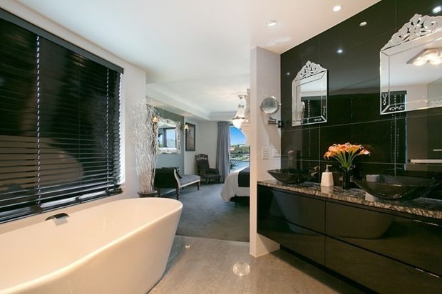 A luxurious contemporary bathroom with a freestanding white bathtub beside large dark horizontal blinds, a glossy black tiled vanity wall, black vessel basins, ornate framed mirrors, and a dark stone benchtop. The bathroom opens directly into a bedroom visible in the background, which contains a bed and upholstered seating. Polished grey flooring and recessed lighting complete the refined interior.