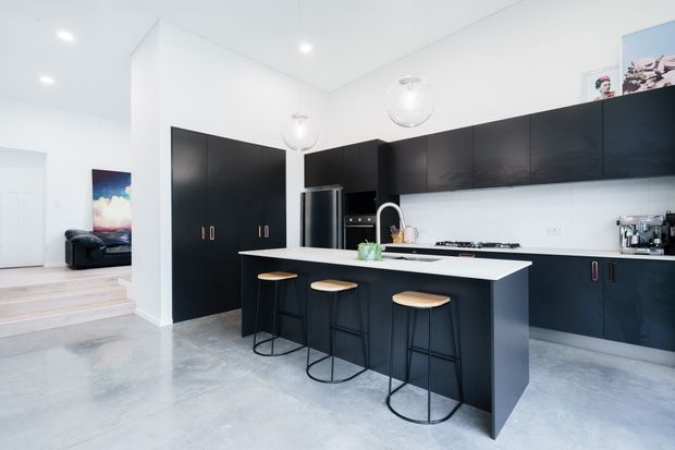 A bright, open-plan contemporary kitchen with extensive matte black cabinetry, a large white island with seating for three, integrated stainless-steel appliances, pendant lights, and a polished grey concrete floor. White walls and ceiling create a crisp, high-contrast interior, with a stepped transition toward an adjacent living area visible at left.