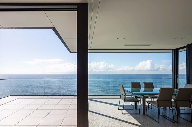 A contemporary open-plan dining and outdoor terrace overlooking the ocean. Full-height glazing and large sliding openings connect the interior dining area to a tiled balcony, with a glass balustrade and uninterrupted sea views. The space features dark structural frames, a pale ceiling with recessed linear ventilation, a glass dining table, and dark upholstered dining chairs.