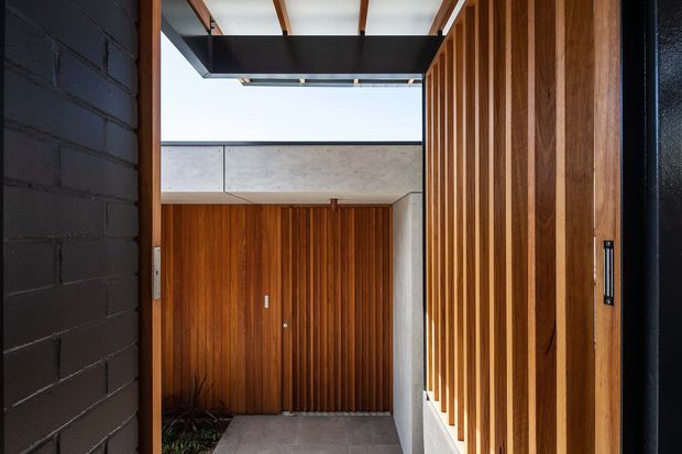 A contemporary residential entry courtyard viewed from a dark interior threshold. The space features warm vertical timber slat screening and a timber-clad entry door, contrasted with exposed concrete panels, dark painted brick, grey tiled paving, and a partially open roof or canopy that brings in daylight. Integrated planting is visible at the base of the timber wall.