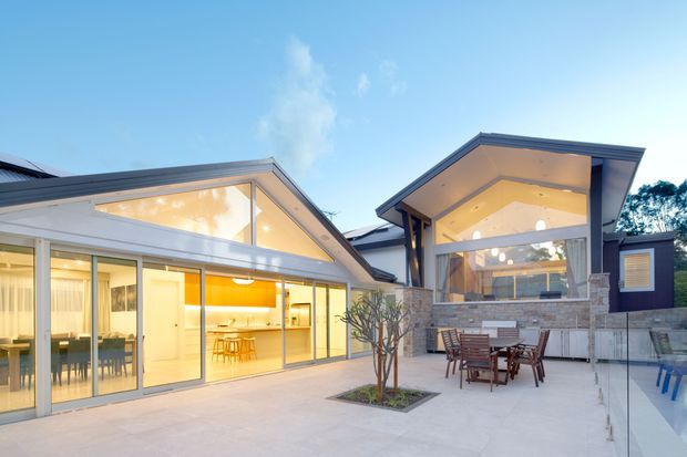Contemporary residential courtyard viewed from outdoors, featuring two connected pavilion-like volumes with steep gabled roofs, extensive floor-to-ceiling glazing, pale stone cladding, and a paved entertaining terrace. Warm interior lighting reveals an open-plan dining and kitchen area, while a dining setting and partial swimming pool are visible in the courtyard.