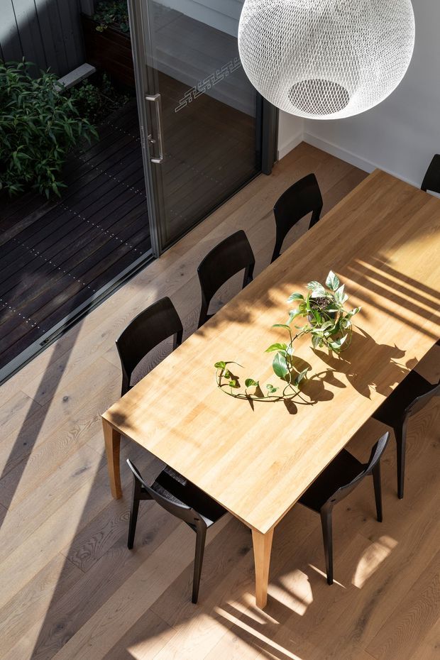 A bright dining area viewed from above, centred on a light timber dining table with dark chairs and a trailing green plant. Pale timber flooring, a large white woven pendant light, and full-height glazed doors connect the room to a dark timber outdoor deck and planted courtyard. Strong sunlight creates pronounced shadows across the table and floor.