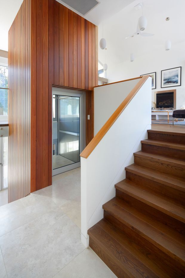 Bright contemporary stairway and landing with a richly stained timber staircase and matching vertical timber wall panelling. A white half-height balustrade with timber handrail separates the stairs from the open living area, while a glazed lift or enclosed opening is visible beside the timber feature wall. The space has pale large-format flooring, white walls and ceiling, pendant lights, a ceiling fan, and glimpses of a living room with a fireplace and framed artwork.