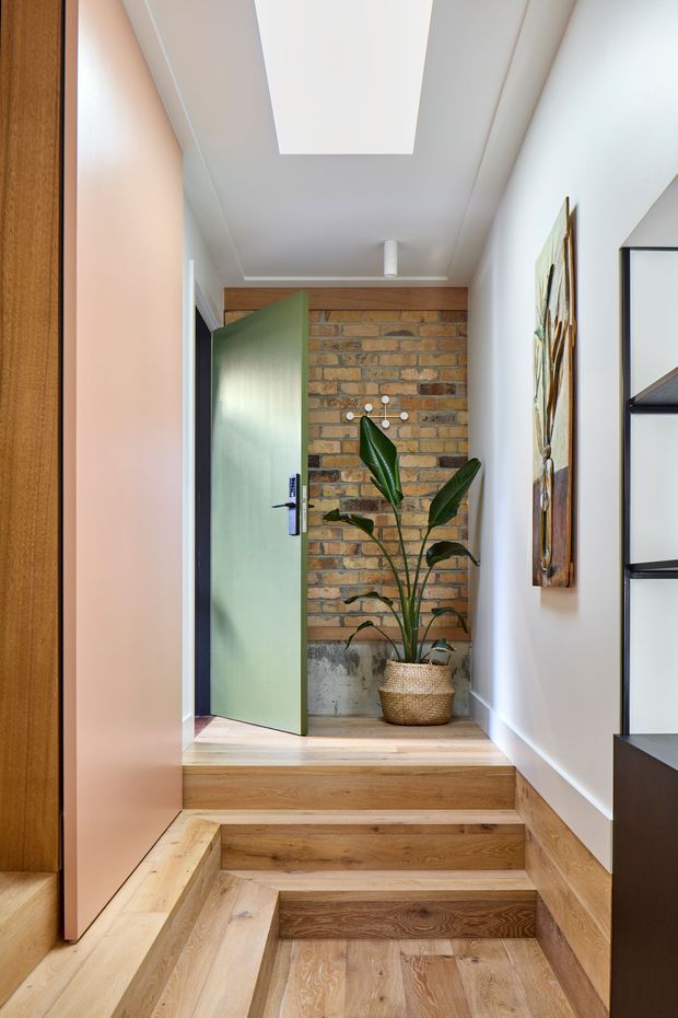 A bright, narrow residential entry hall with a short set of timber-clad steps leading up to an open pale green door. The rear wall features exposed warm brick with a lower concrete section, while white walls and ceiling surround a large rectangular skylight. A potted tropical plant, wall artwork, and dark built-in shelving add detail to the warm timber interior.