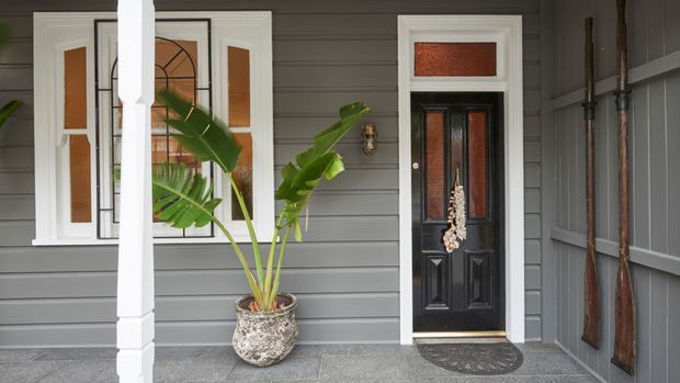 Covered residential front porch with grey horizontal weatherboard cladding, white trim and columns, a black decorative glazed entry door, leadlight-style window, and a potted tropical plant. Vertical timber paddles or boards are mounted along the right wall.